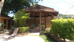 A covered outdoor seating area with a tiled roof, surrounded by greenery and a pathway leading to a building entrance. - Villa in a Quiet and Safe Residential Area for 6 people. (Comunidad, Carrillo)