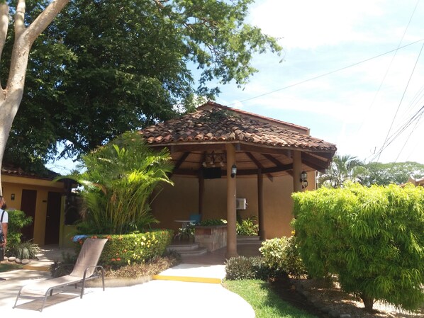 A covered outdoor seating area with a tiled roof, surrounded by greenery and a pathway leading to a building entrance.