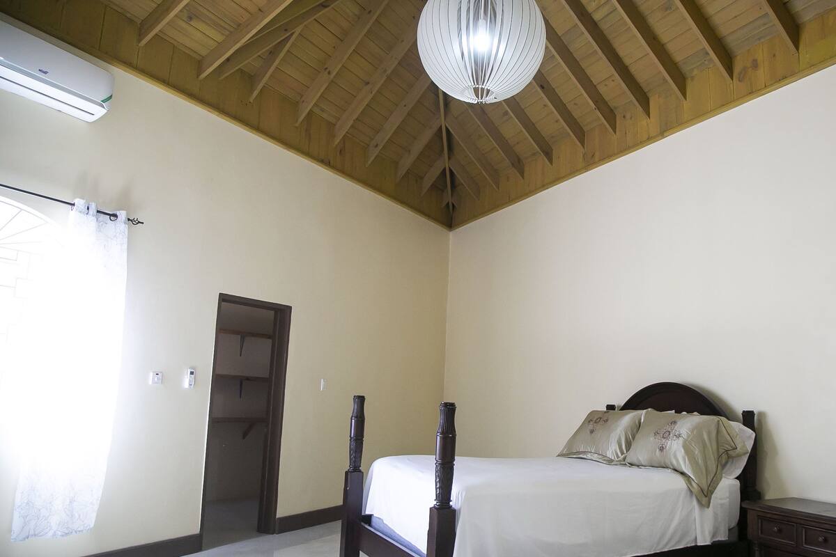A bedroom with a wooden ceiling, a bed with a headboard, a nightstand, and an air conditioning unit.