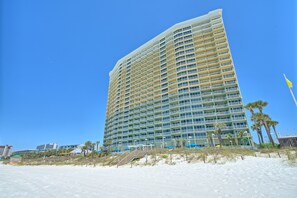Un grand hôtel à plusieurs étages, doté de balcons, situé près d’une plage de sable bordée de palmiers.