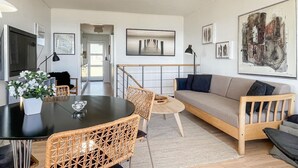 A modern living room with a sofa, a dining table, and chairs. There is a TV, artwork on the walls, and a staircase leading to another room.