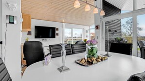 Een moderne eetruimte met een grote tafel, zwarte lederen stoelen, een aan de muur bevestigde televisie en uitzicht op het omliggende landschap door grote ramen.