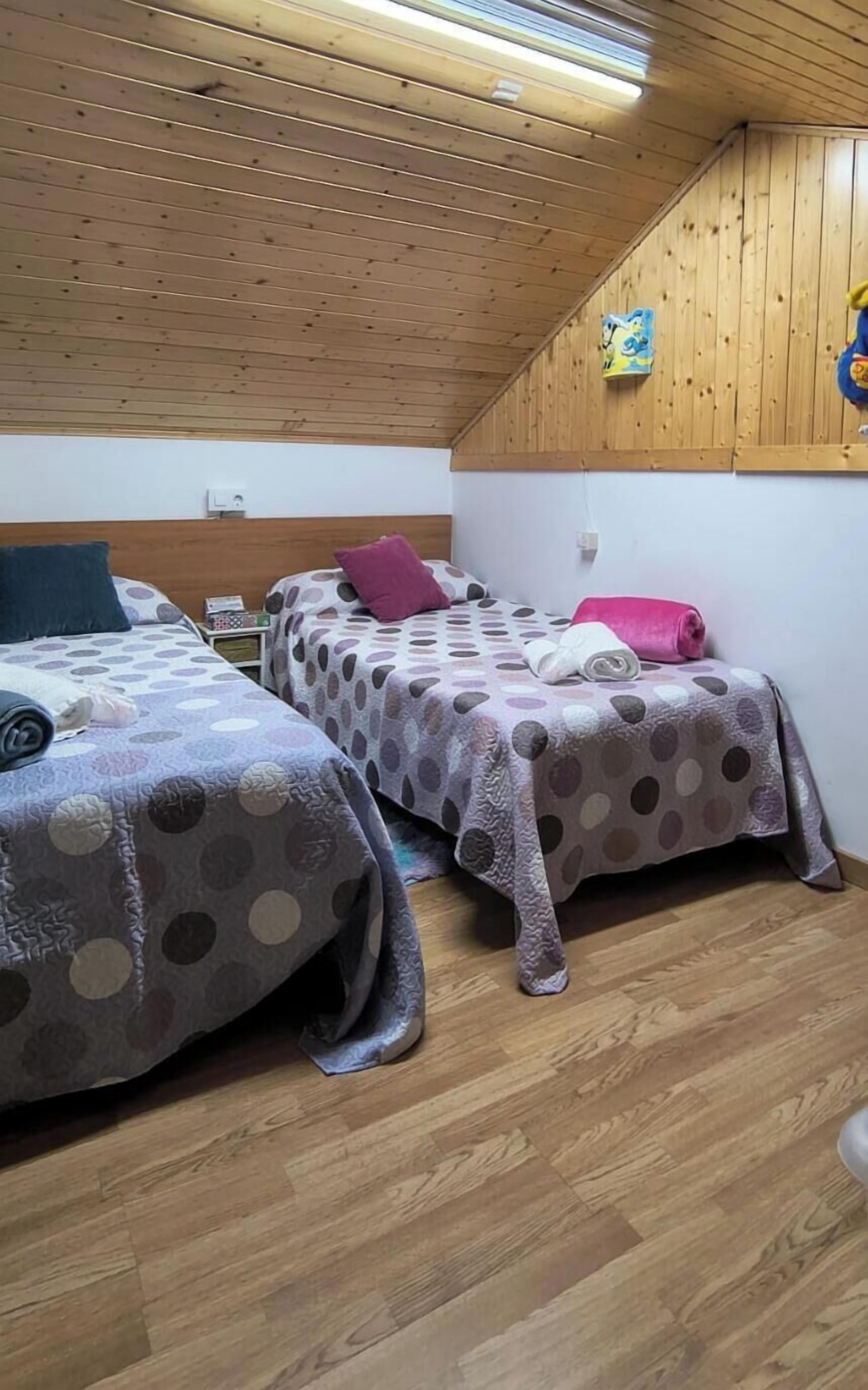 A room with two beds, wooden flooring, a sloped ceiling, and a wall-mounted light fixture.