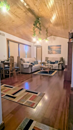 A spacious living room with wooden flooring, a sofa set, a dining area with chairs, and a painting on the wall.