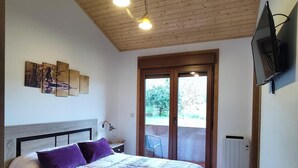 A bedroom with a wooden ceiling, a bed with a patterned bedspread, a desk with a lamp, and a TV mounted on the wall.