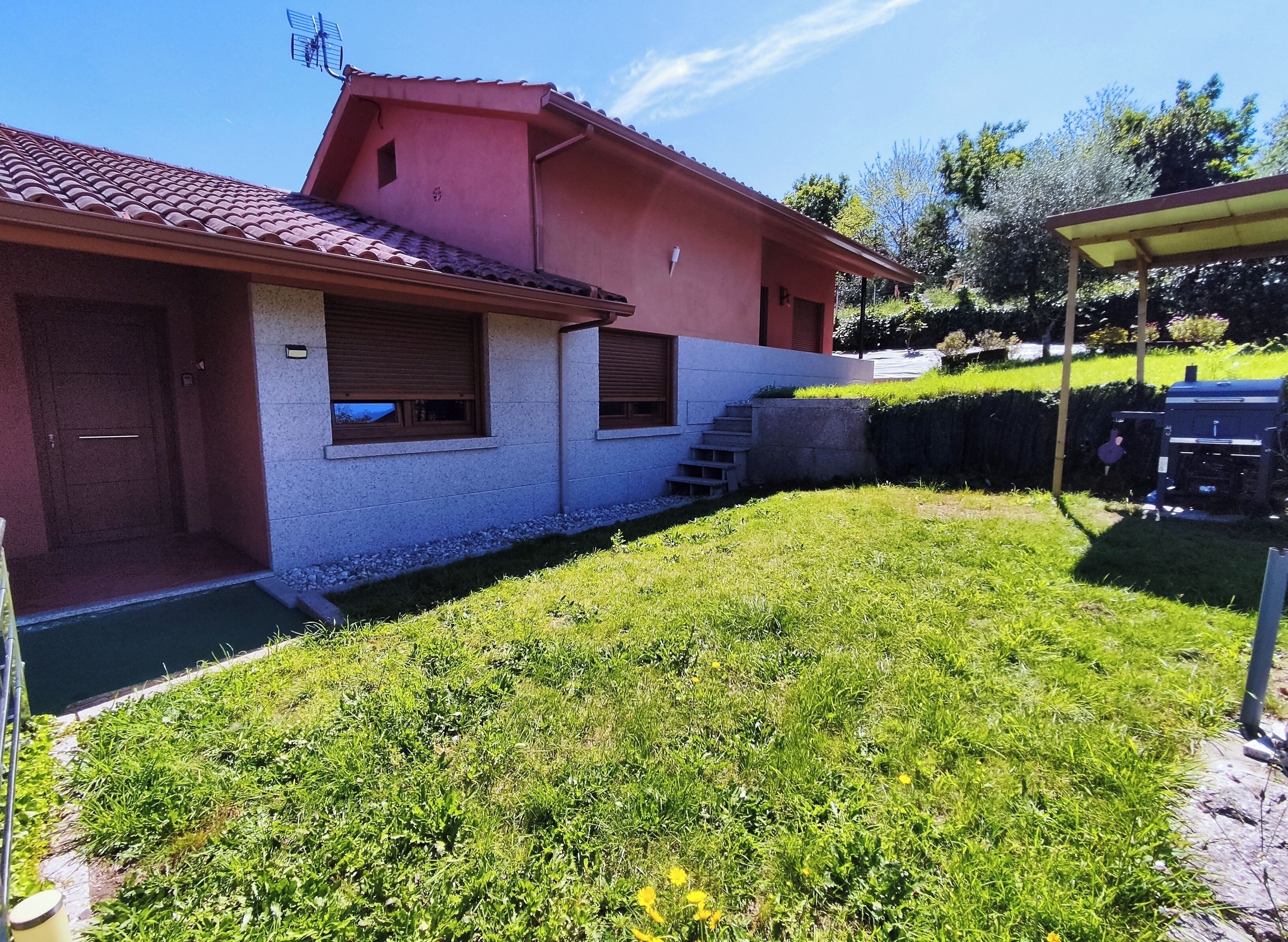 Una casa de dos pisos con techo rojo y paredes blancas, un pequeño jardín delantero y un espacio exterior cubierto.