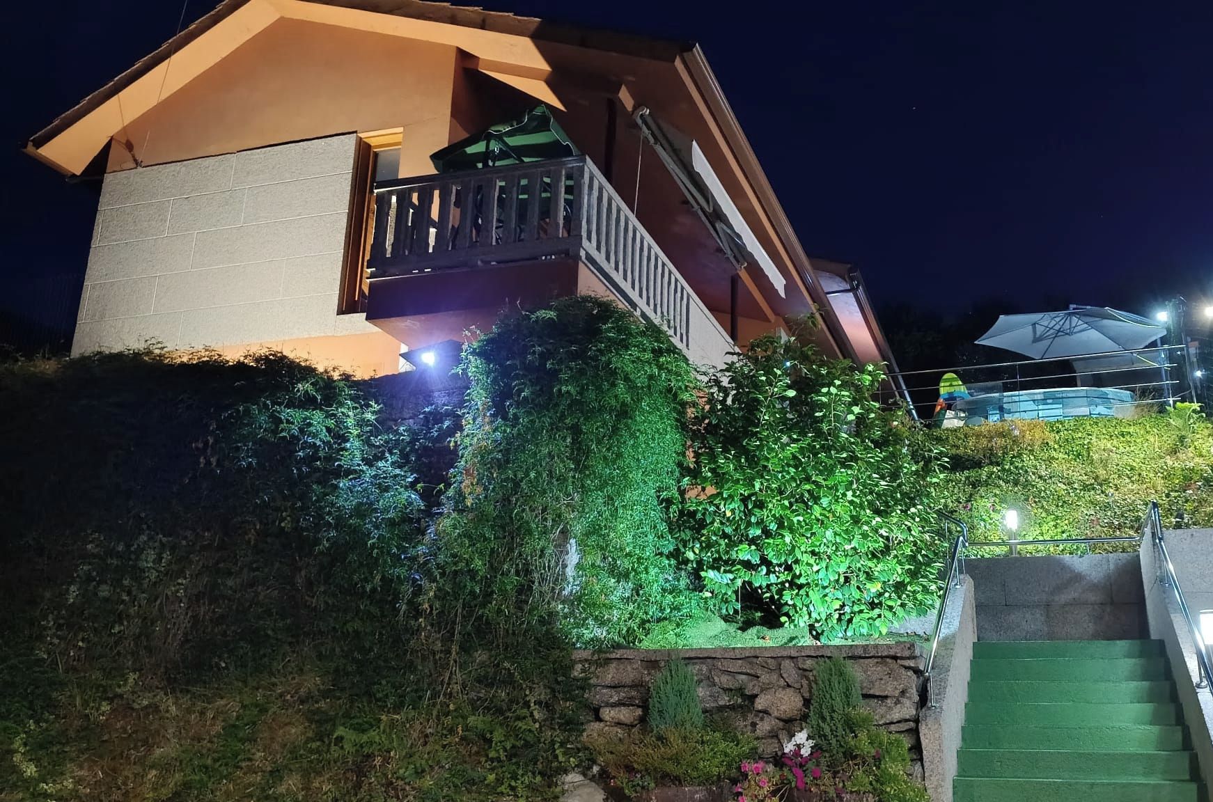 Una casa moderna con balcón, rodeada de vegetación exuberante y un muro de piedra.