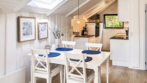 Un espace repas avec une table et des chaises blanches, des œuvres d’art encadrées et un luminaire suspendu.