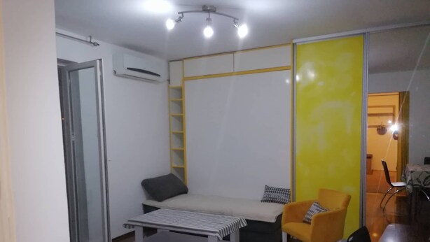 Une pièce de taille modeste, comprenant un lit, une petite table, une chaise et un mur de séparation jaune.