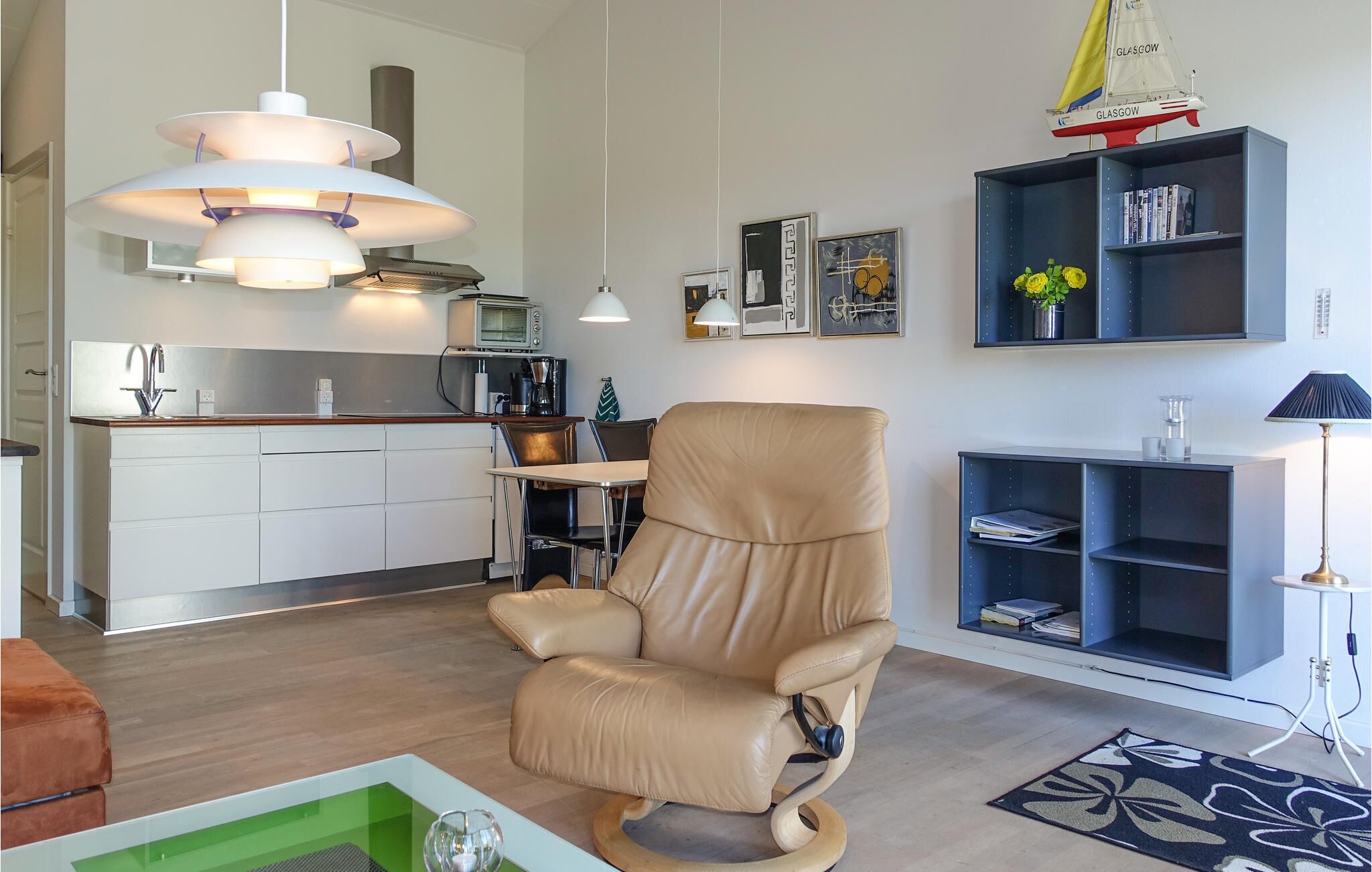 Una sala moderna con un sillón reclinable de cuero, un comedor con mesa y sillas, una cocina con un tostador y una estantería con libros y una planta.