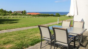 An outdoor patio with a table and chairs, a view of the sea, and a white building in the background. - Nice apartment in Allinge with WiFi (Allinge)