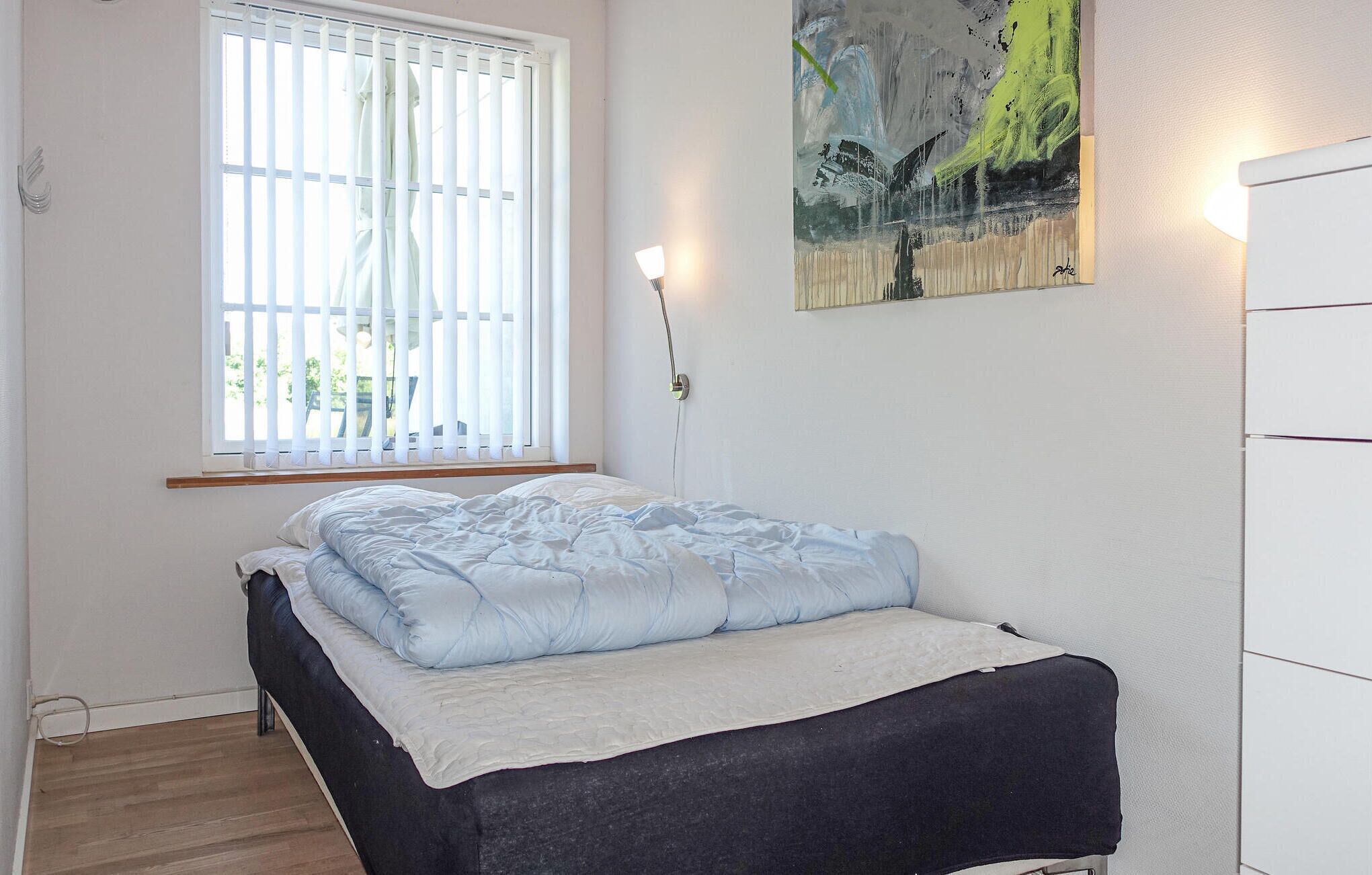 Une chambre à coucher avec un lit, une fenêtre avec des stores, un tableau au mur et une lampe de chevet.