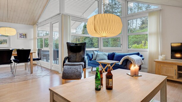 A spacious living room with a large window, a blue sofa, a wooden table with bottles, and a TV on a wooden cabinet. - 2 bedroom lovely home in Aabenraa (Aabenraa)