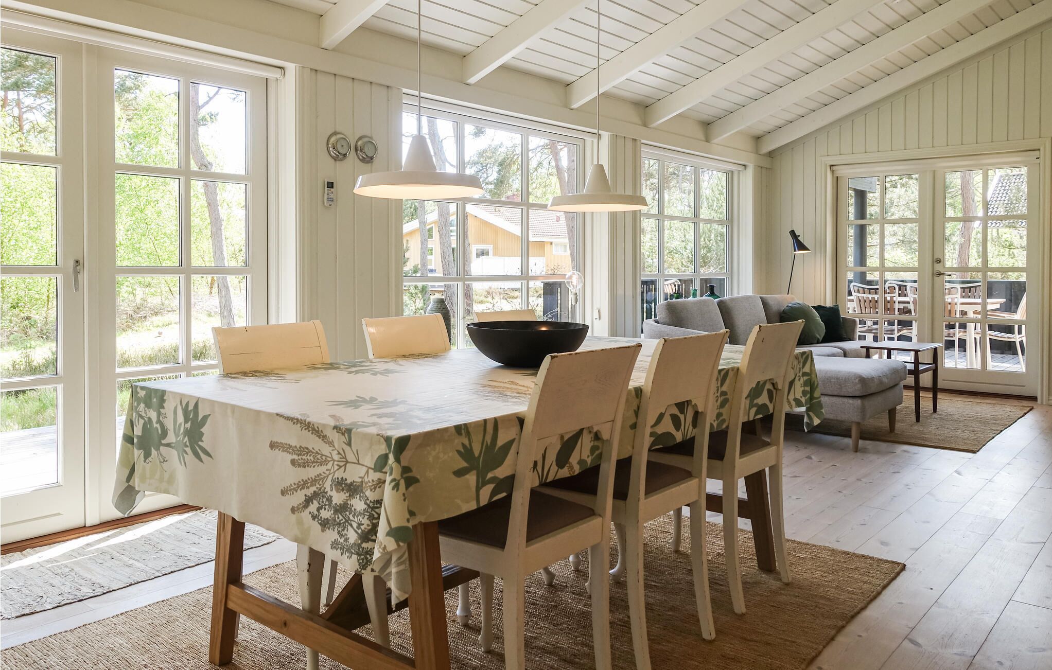 Un coin repas avec une table dressée pour quatre personnes, entourée de chaises à l’assise recouverte d’un tissu à motifs floraux. Deux lampes suspendues éclairent la table, et une grande fenêtre offre une vue sur l’extérieur.