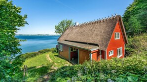 Een rieten huis met een rode gevel, gelegen aan een waterpartij, omringd door groen.