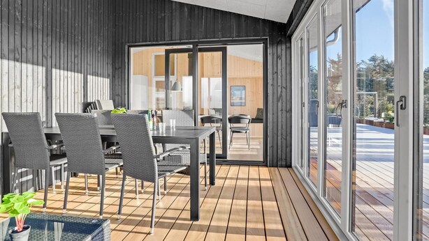 Ett modernt utomhusområde för måltider med ett trädäck, stolar med metallram och en glasdörr som leder in till ett inomhusutrymme.