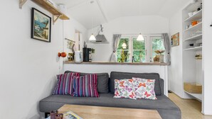 A cozy living room with a grey sofa, striped pillows, and a wooden coffee table. A window with curtains and a view of greenery is visible. - Nice home in Nexø with kitchen (Nexø)