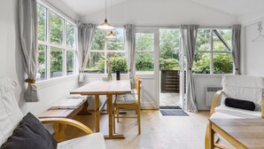 A bright, open-plan room with a wooden dining table, chairs, and a bench. Large windows provide views of greenery outside. - Nice home in Nexø with kitchen (Nexø)