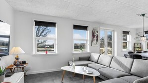 Ett modernt vardagsrum med en grå soffa, ett vitt soffbord och stora fönster som ger utsikt över omgivningen.
