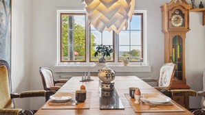 A dining room with a wooden table set for two, a large window with a view of greenery, a grandfather clock, and a decorative hanging light fixture. - Nice home in Nexø with WiFi (Nexø)