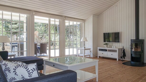 A modern living room with a glass coffee table, a sofa with patterned cushions, a TV, a fireplace, and large windows offering a view of trees. - 3 bedroom gorgeous home in Nexø (Nexø)