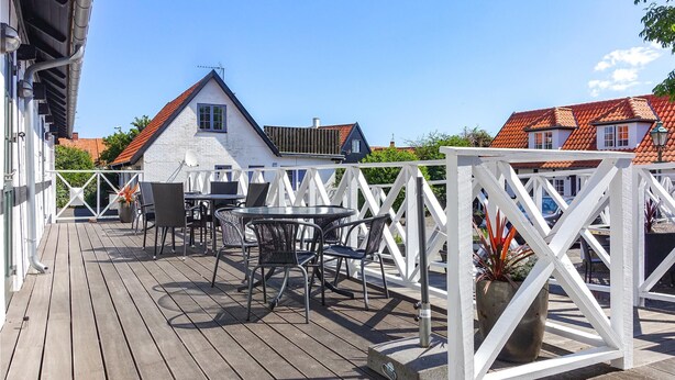 Une terrasse en bois avec des sièges d’extérieur, une clôture blanche et, au fond, des bâtiments aux toits recouverts de tuiles rouges.