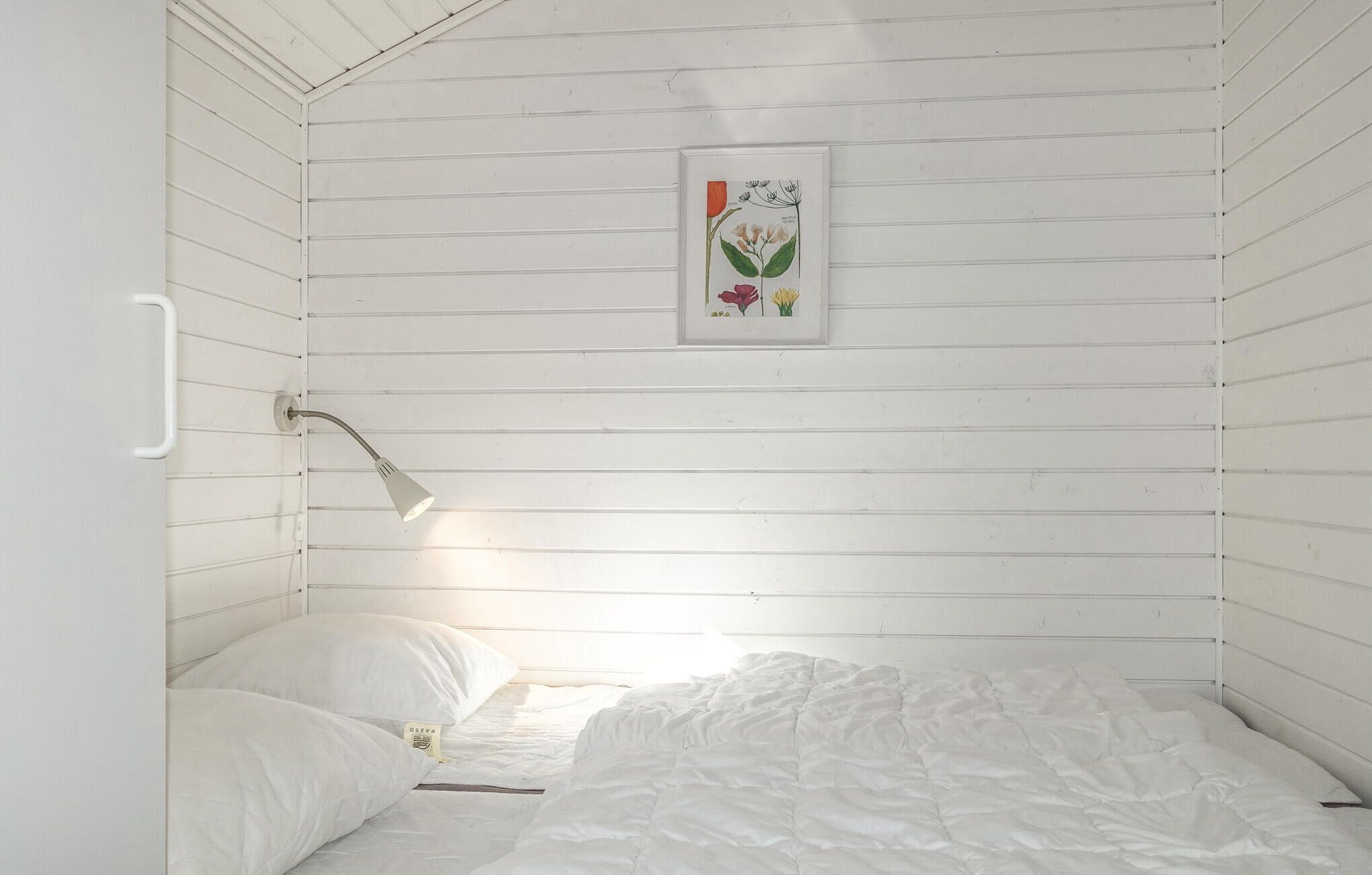 A small, white, wooden room with a single bed, a bedside lamp, and a framed botanical illustration on the wall.