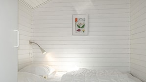 A small, white, wooden room with a single bed, a bedside lamp, and a framed botanical illustration on the wall.