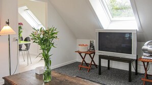 A cozy attic room with a sloped ceiling, a television on a stand, a vase with flowers, and a lamp. - 2 bedroom nice apartment in Gudhjem (Gudhjem)