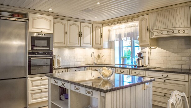 Una cocina con armarios blancos, una isla central, electrodomésticos de acero inoxidable y un ventanal con cortinas.