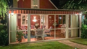Un área de comedor al aire libre techada, con sillas de mimbre y una mesa, rodeada de puertas y ventanas de vidrio.