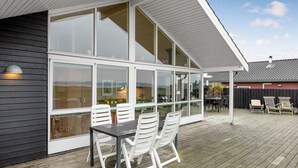 Een overdekt terras met een tafel en stoelen, grote ramen en uitzicht op de achtertuin.
