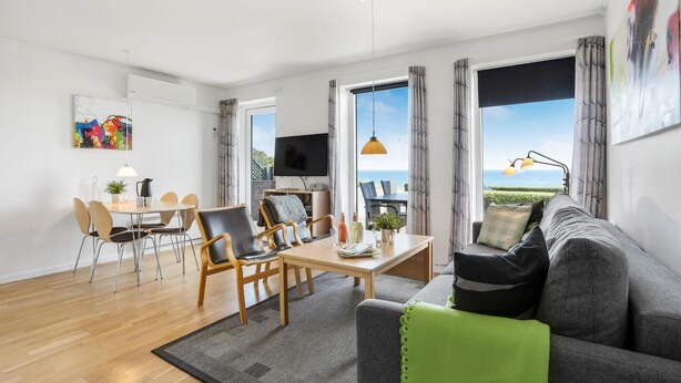 Una sala de estar moderna con sofá, mesa de comedor y sillas. Hay un televisor y una ventana con vistas al mar.