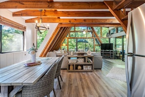 A modern kitchen with a dining area, wooden beams, large windows, and a built-in entertainment unit. - Lakefront A-Frame · Hot Tub · Paddle Pontoon Boat (Innsbrook)