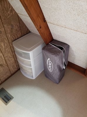 Ein Raumeck mit einem weißen Kunststoffregal und einer grauen GRACO-Tasche an einer strukturierten Wand und einem Holzbalken.