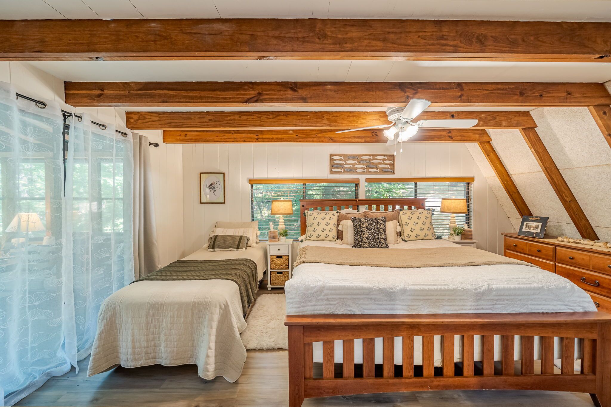A bedroom with a large bed, two smaller beds, wooden beams, a ceiling fan, and a window with blinds.
