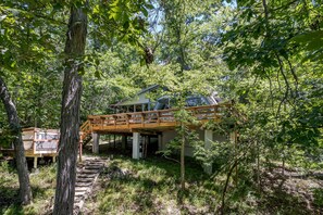 A cabin with a wooden deck and a staircase leading to a second-story balcony, surrounded by dense forest. - Lakefront A-Frame · Hot Tub · Paddle Pontoon Boat (Innsbrook)