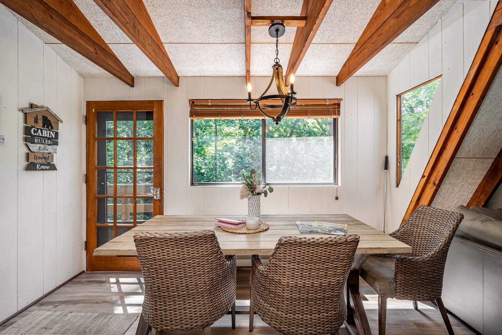 A dining area with a wooden table, wicker chairs, a chandelier, and a window with a view of trees.