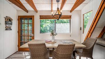 A dining area with a wooden table, wicker chairs, a chandelier, and a window with a view of trees.
