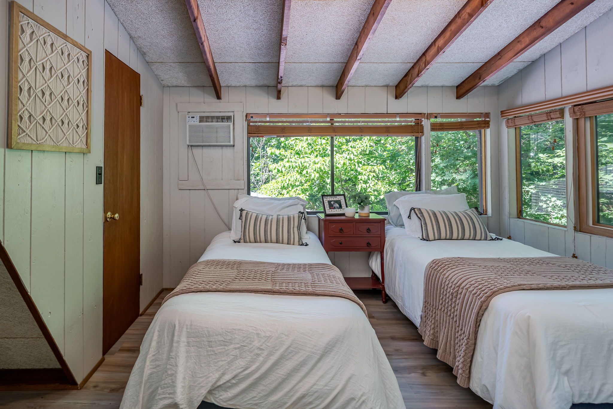 A bedroom with two beds, a wooden door, a window with a view of greenery, and a wall-mounted air conditioning unit.