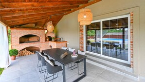 Un patio exterior con un horno de ladrillo, una mesa de comedor con sillas y un ventanal con vistas a una piscina.