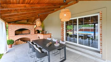 Eine Terrasse im Freien mit einem Backofen aus Ziegelsteinen, einem Esstisch mit Stühlen und einem großen Fenster mit Blick auf einen Pool.