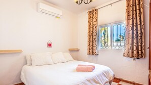 A bedroom with a bed, white curtains, a window with a view, and an air conditioning unit.
