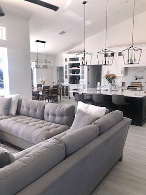 Un soggiorno moderno con un ampio divano a forma di L grigio, un'area pranzo con un mobile bianco e lampade a sospensione, e una cucina con pensili bianchi e un piano di lavoro in marmo.