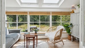 Ein Wintergarten mit großen Fenstern, einem Holztisch, einer Couch und einem Sessel.