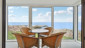 Ein Zimmer mit einem großen Fenster mit Blick auf eine felsige Küstenlinie und das Meer. Ein runder Tisch mit vier Korbstühlen.