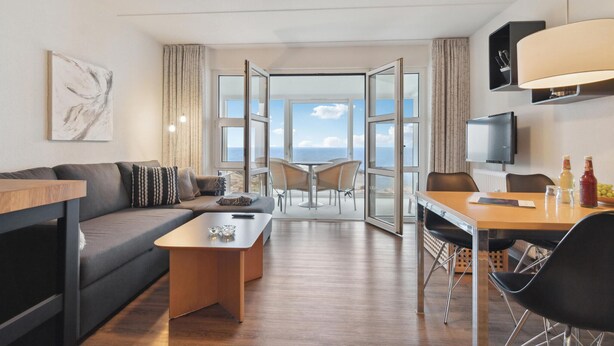 Ein modernes Hotelzimmer mit einer Couch, einem Couchtisch, einem Esstisch mit Stühlen, einem Fernseher und Meerblick durch große Fenster.