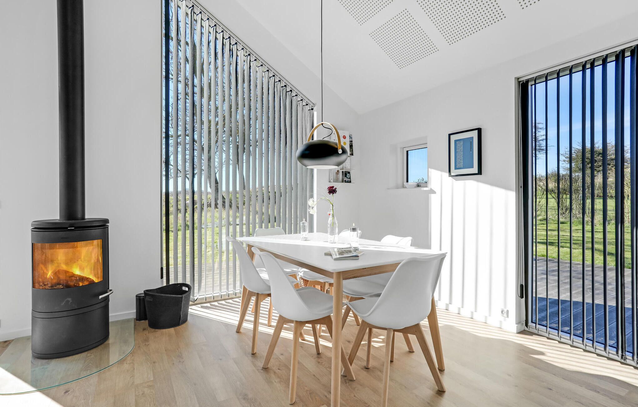 A modern room with a wood-burning stove, a dining table with chairs, and large windows with vertical blinds.