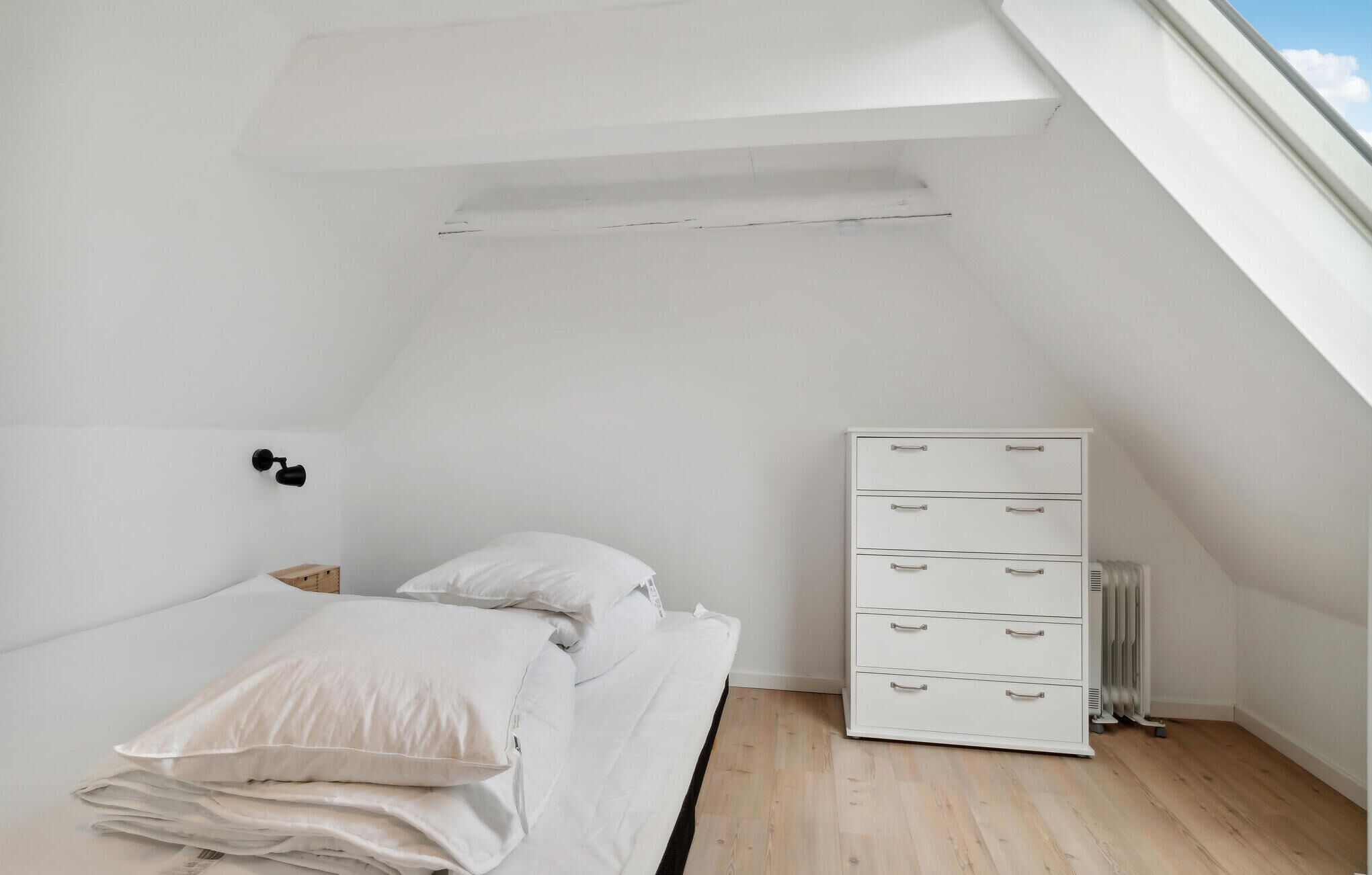 A small, white bedroom with a sloped ceiling, a bed with white bedding, a white dresser with drawers, and a radiator.