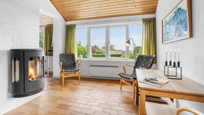 Une pièce chaleureuse avec une cheminée, deux fauteuils, une table et une grande fenêtre avec des rideaux.